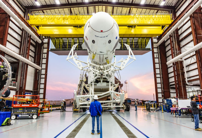 A man in a hardhat stands before a giant rocket in an even larger hanger.