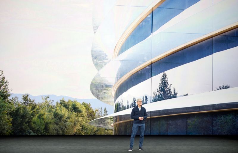 A hoody man stands before a multi-story glass building.