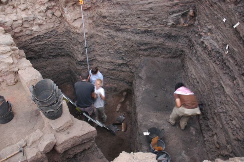 Color photo of archaeologists excavating layers of copper production waste in a deep pit.