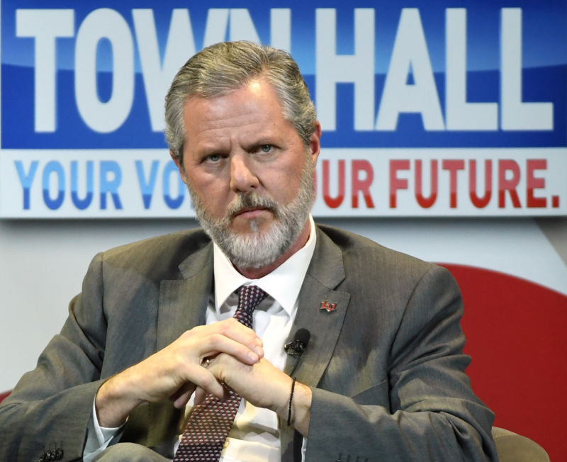 A serious man in a suit sits on a stage.