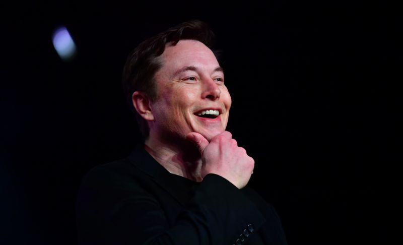 A man laughs while scratching his chin against an indistinct dark background.