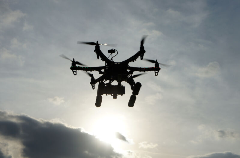 A drone in flight is silhouetted against a cloudy sky.