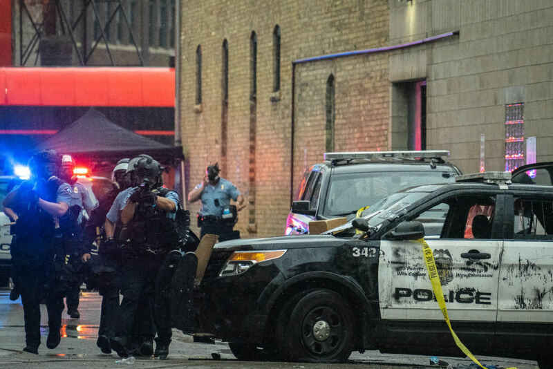 Police officers in riot gear advance, fangs out, down a city street.