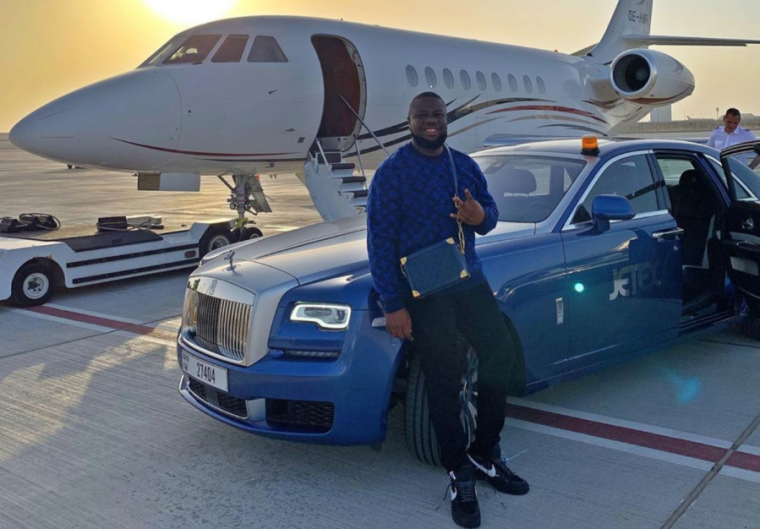 A smiling man leans against an expensive car parked in front of a private jet.