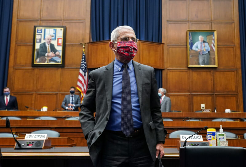 A man in a suit and a face mask stands in a wood-paneled room.