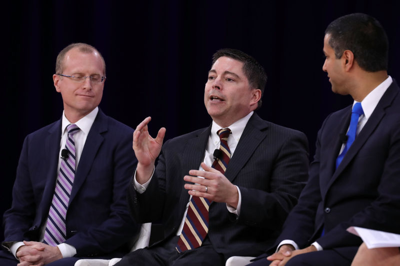 FCC Commissioner Michael O'Rielly speaks at a conference while FCC Commissioner Brendan Carr and Chairman Ajit Pai look on.