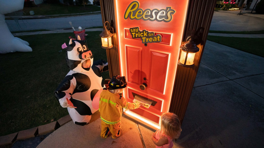 An orange door that says Reese's say trick or treat with a kid dressed as a firefighter and a cow and a ballerina taking something from the door's mail slot