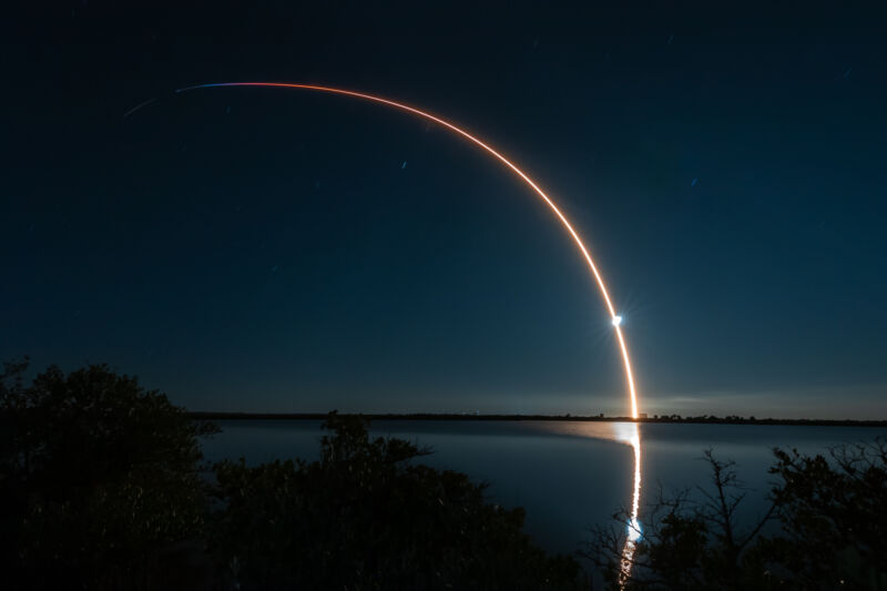 A streak of light arcs across the night sky and is reflected in a lake.