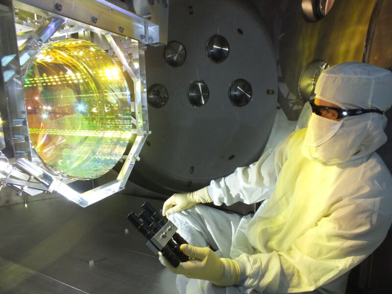 A researcher in protective gear examines an impossibly futuristic mirror.