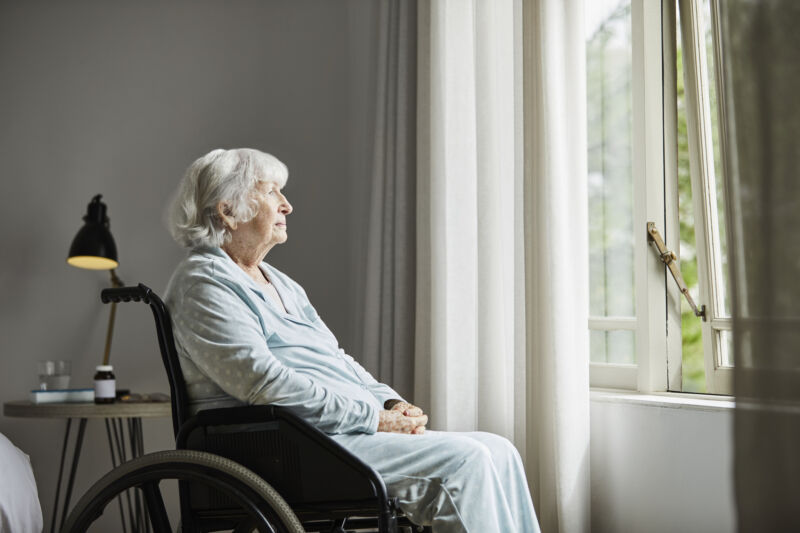 A white-haired woman in a wheelchair looks out a window.
