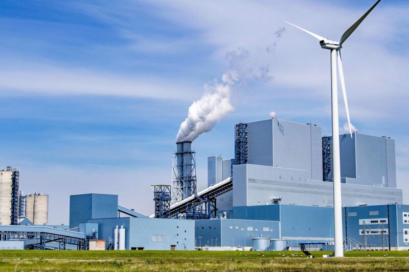 Steam rises from an industrial facility behind a wind turbine.