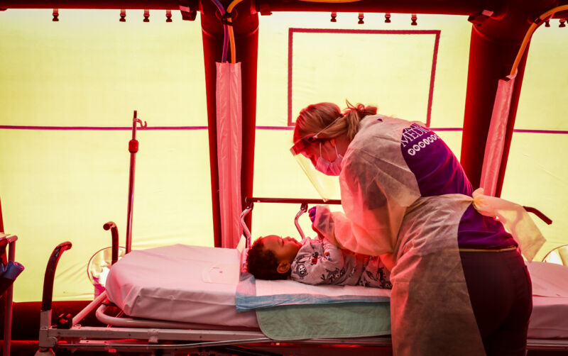 A woman in protective gear leans over a toddler in a bed.