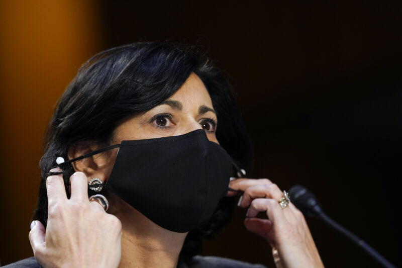 A woman adjusts her face mask while sitting in front of a microphone.