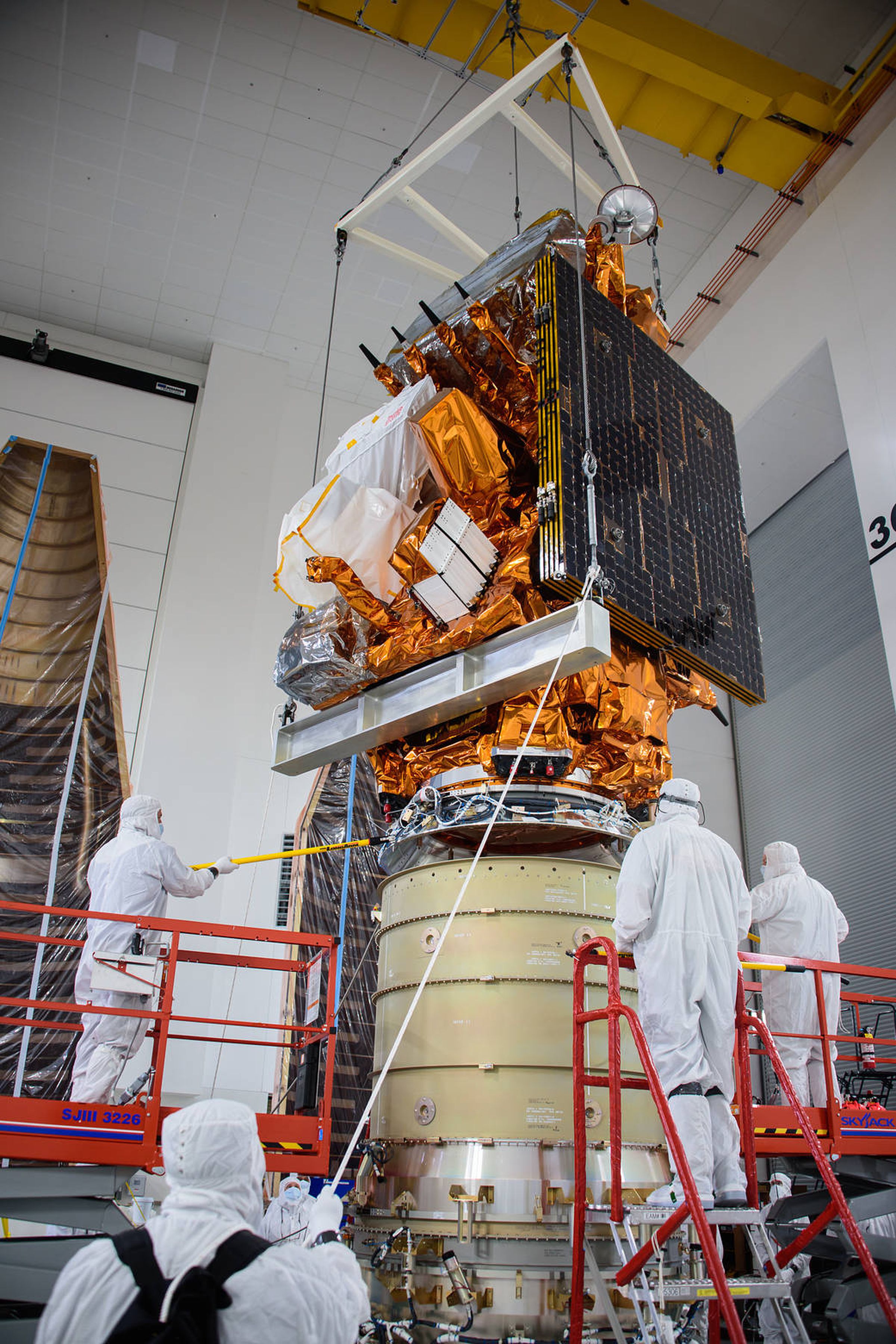 four people in white suits stand in a clean room around a satellite