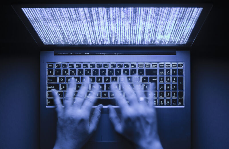 Blurred hands are typing on a laptop computer in the dark with illuminated keyboard and illegible mystic program code visible on the screen.