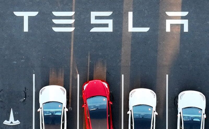 Aerial view of cars parked in a Tesla factory parking lot. A Tesla logo is painted on the concrete.