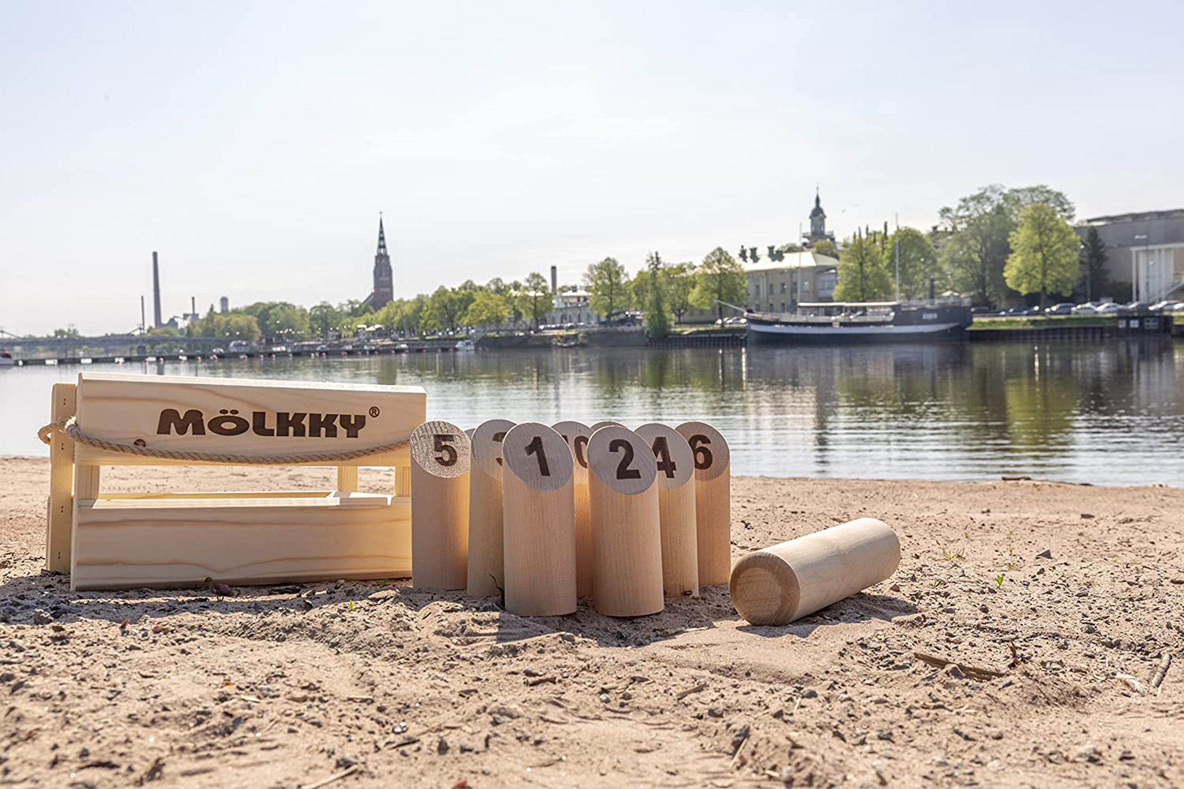 Molkky game with a wooden, several numbered pins, and a wooden dowel, on a beach with a town in the background.