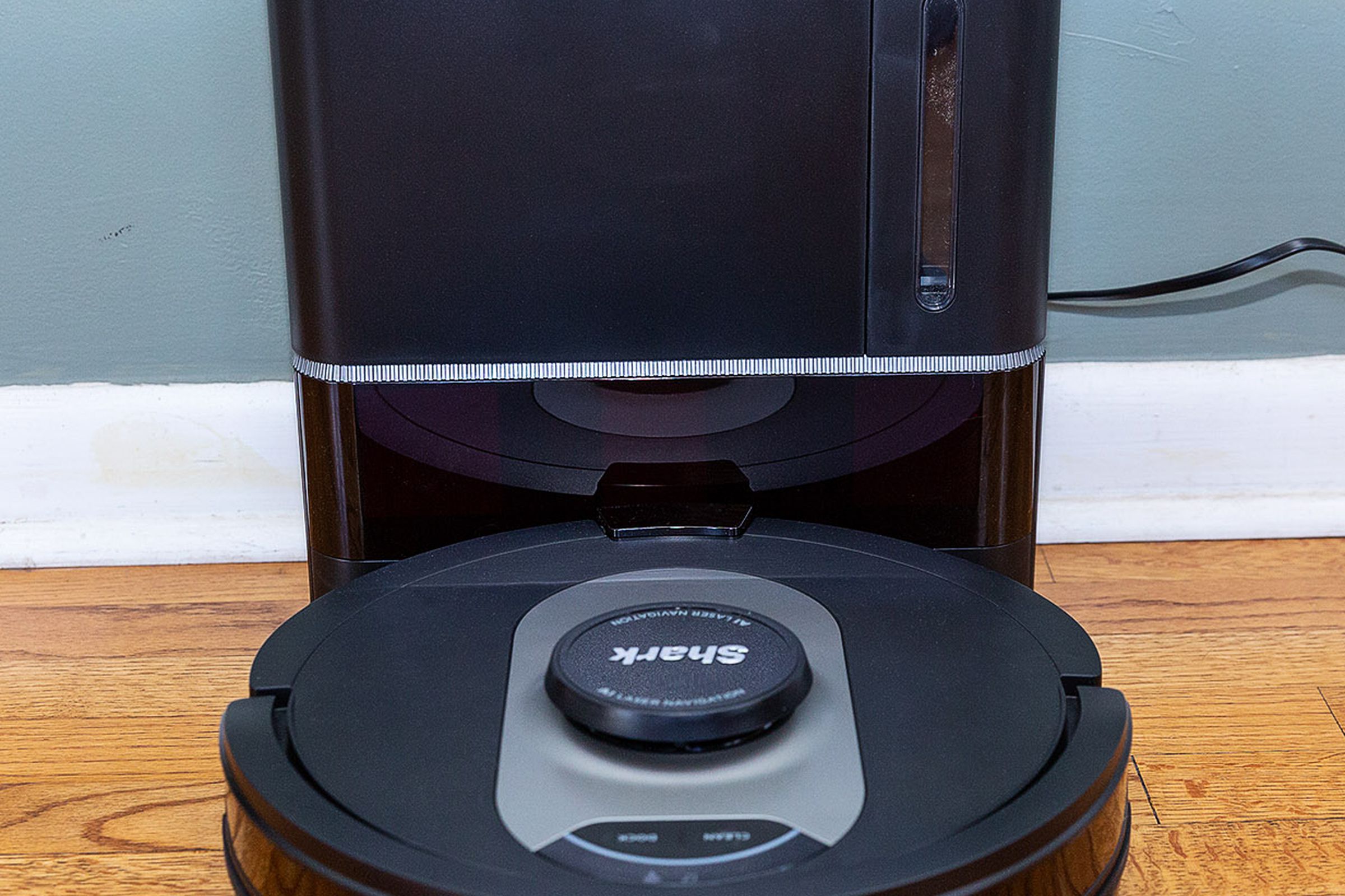 A head-on angle of the Shark AV2501AE AI Robot Vacuum docked in its self-cleaning station against a wall, plugged into a nearby outlet.