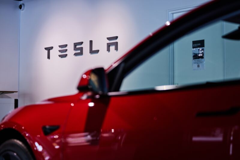A Tesla car is viewed from the side inside a store. A Tesla logo is on the wall behind the car.