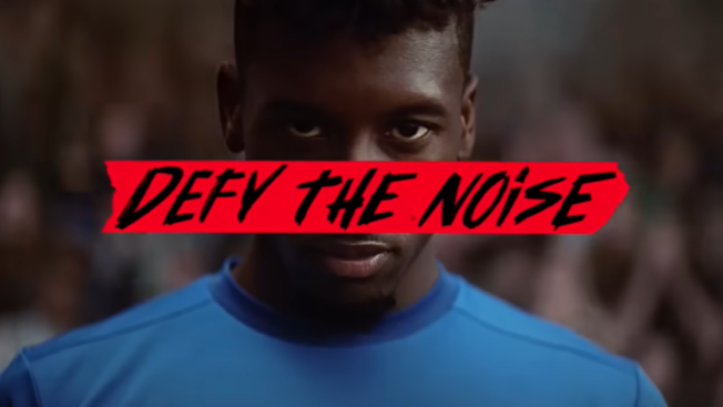 A close-up of a soccer player's face with the words "Defy the Noise" written on a red banner and placed over his face