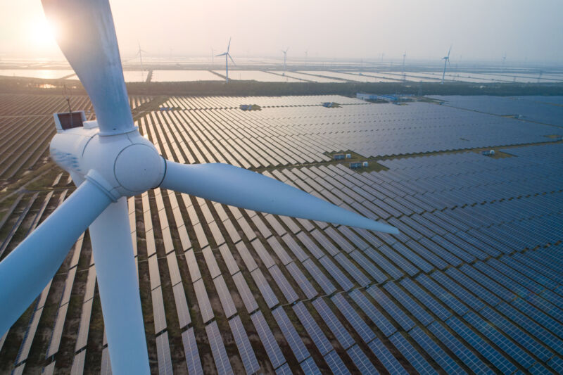 Wind turbines stand above a large field of solar panels in a view backlit by a rising Sun.
