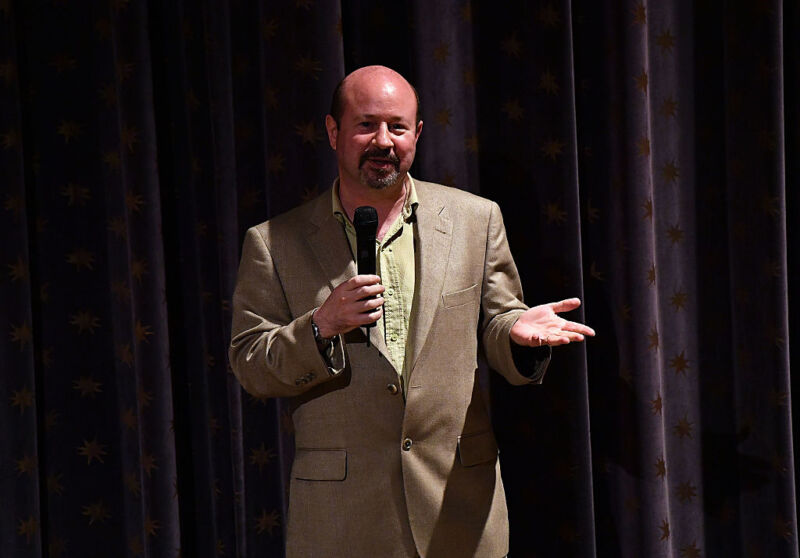 Image of a middle-aged male speaking into a microphone against a dark backdrop.