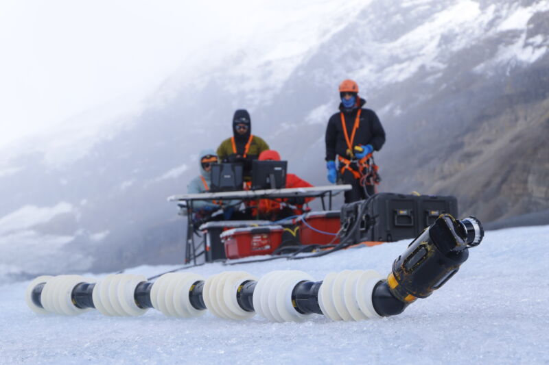 Image of two humans sitting behind a control console dressed in heavy clothing, while a long tube sits on the ice in front of them.