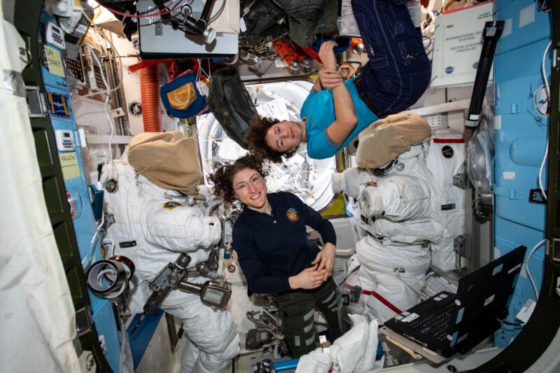 Image of two astronauts in an equipment filled chamber, standing near the suits they wear for extravehicular activities.