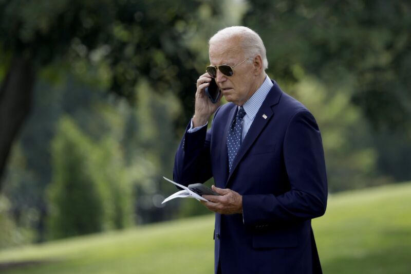 President Biden walking outdoors while holding a cell phone to his ear with one hand and holding another phone in his other hand.