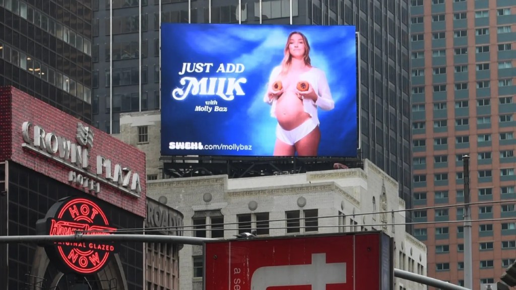 Bright blue digital billboard in Times Square showing woman holding cookies in front of her breasts while wearing white button down shirt and underwear. Text reads 'Just Add Milk with Molly Baz.'