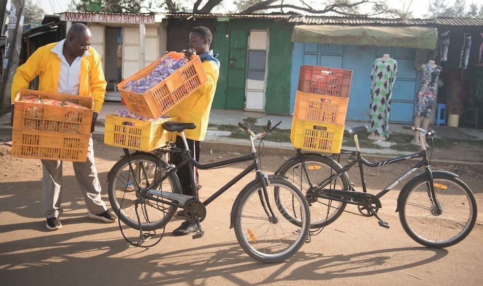 Two people loading yellow milk-style crates of cargo onto Buffalo bicycles, seemingly in the street of a small village.