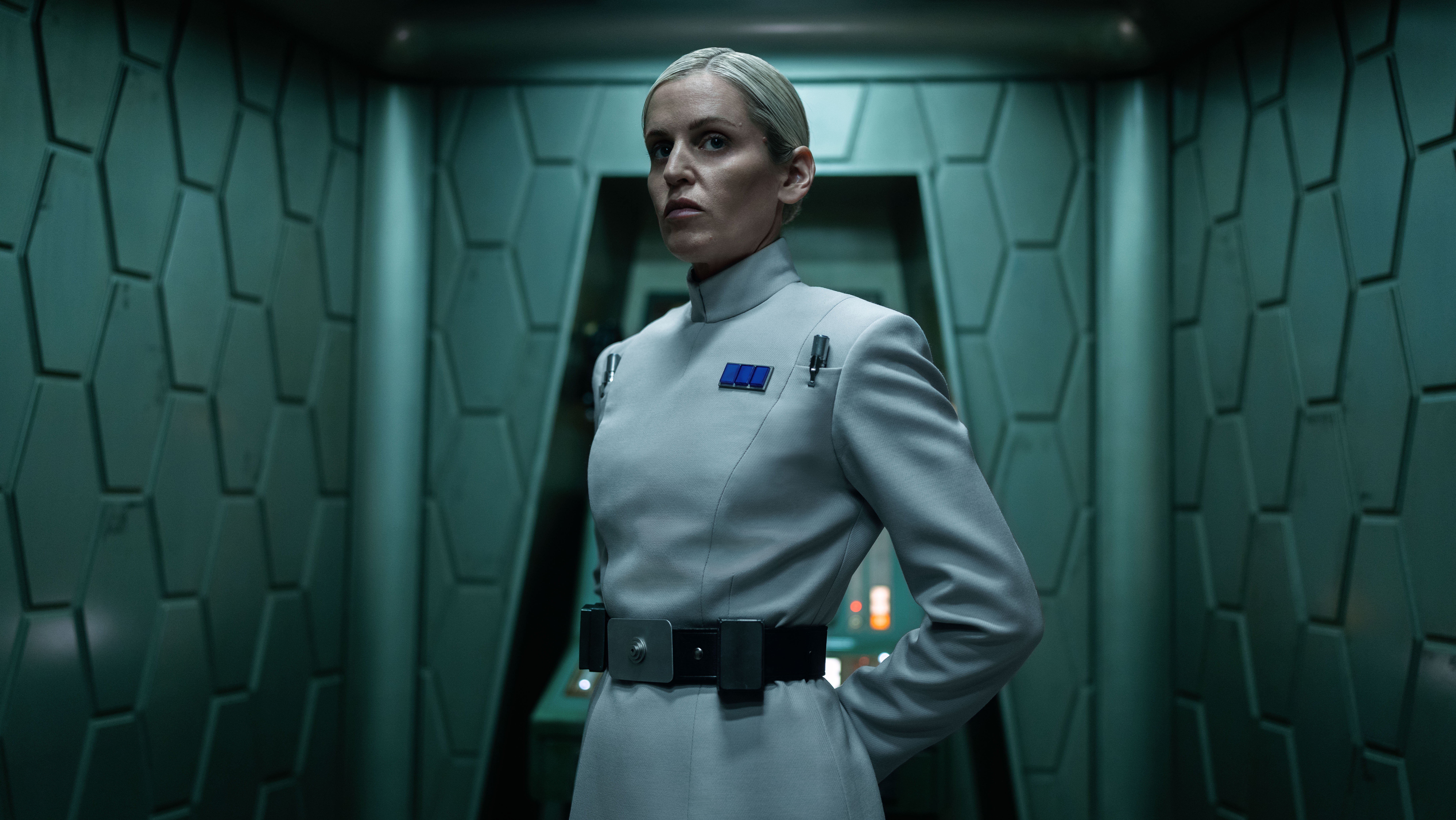 A woman in a white coat with a black utility belt. The woman is standing with her arms behind her back in a dimly lit room whose walls are lined with metallic hexagons.