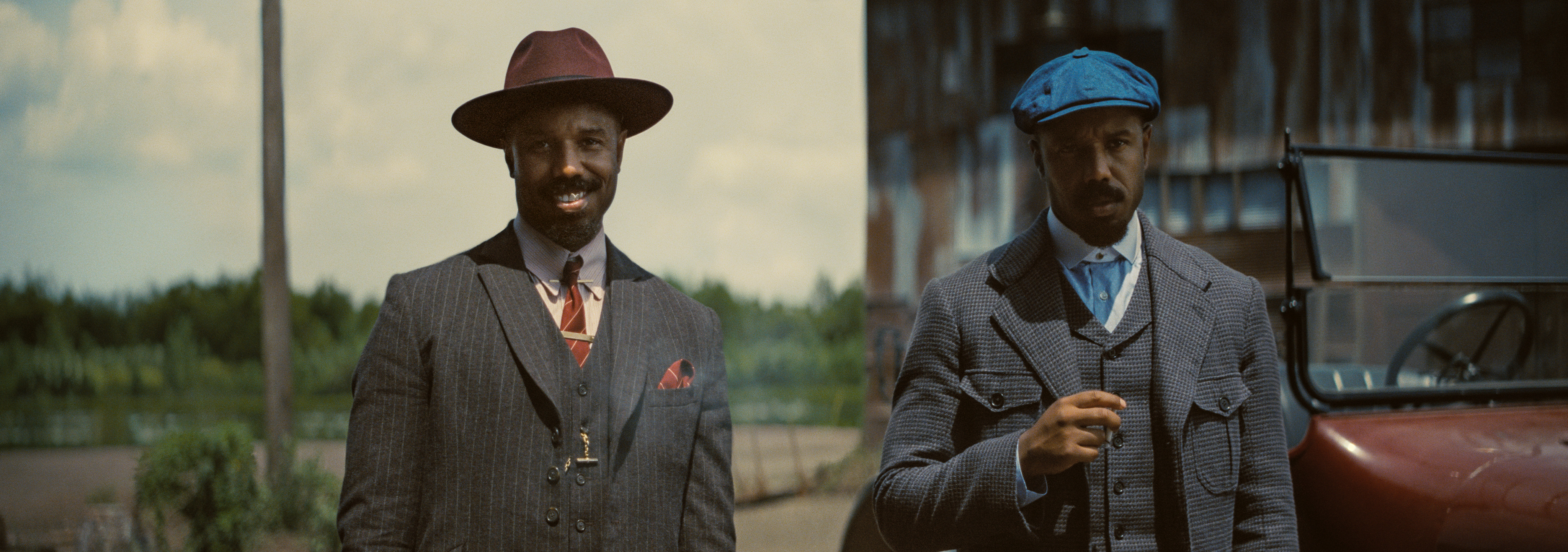 A pair of men who are twins wearing similar but slightly different gray suits. The man on the left is wearing a burgundy fedora while the man on the right is wearing a blue page boy hat.