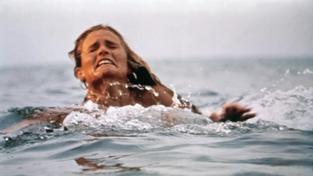 closeup of woman's face and head just above the water's surface, in agony as an unseen shark drags her back and forth