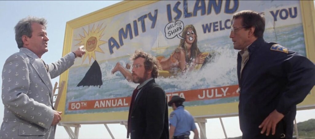 Bearded man standing in front of a vandalized billboard, showing a female surfer being pursued by a visible shark fin sticking out of the water