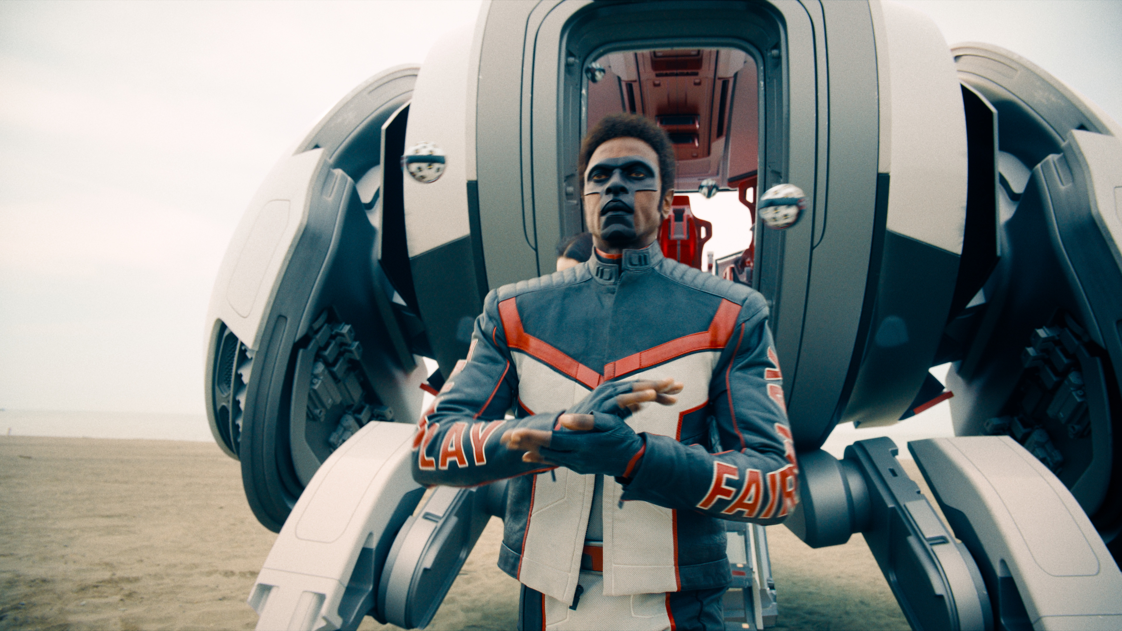 A man with a black face mask shaped like the letter T. The man is also a black, red, and white motorcycle jacket with matching pants and standing in front of a pherical space ship.