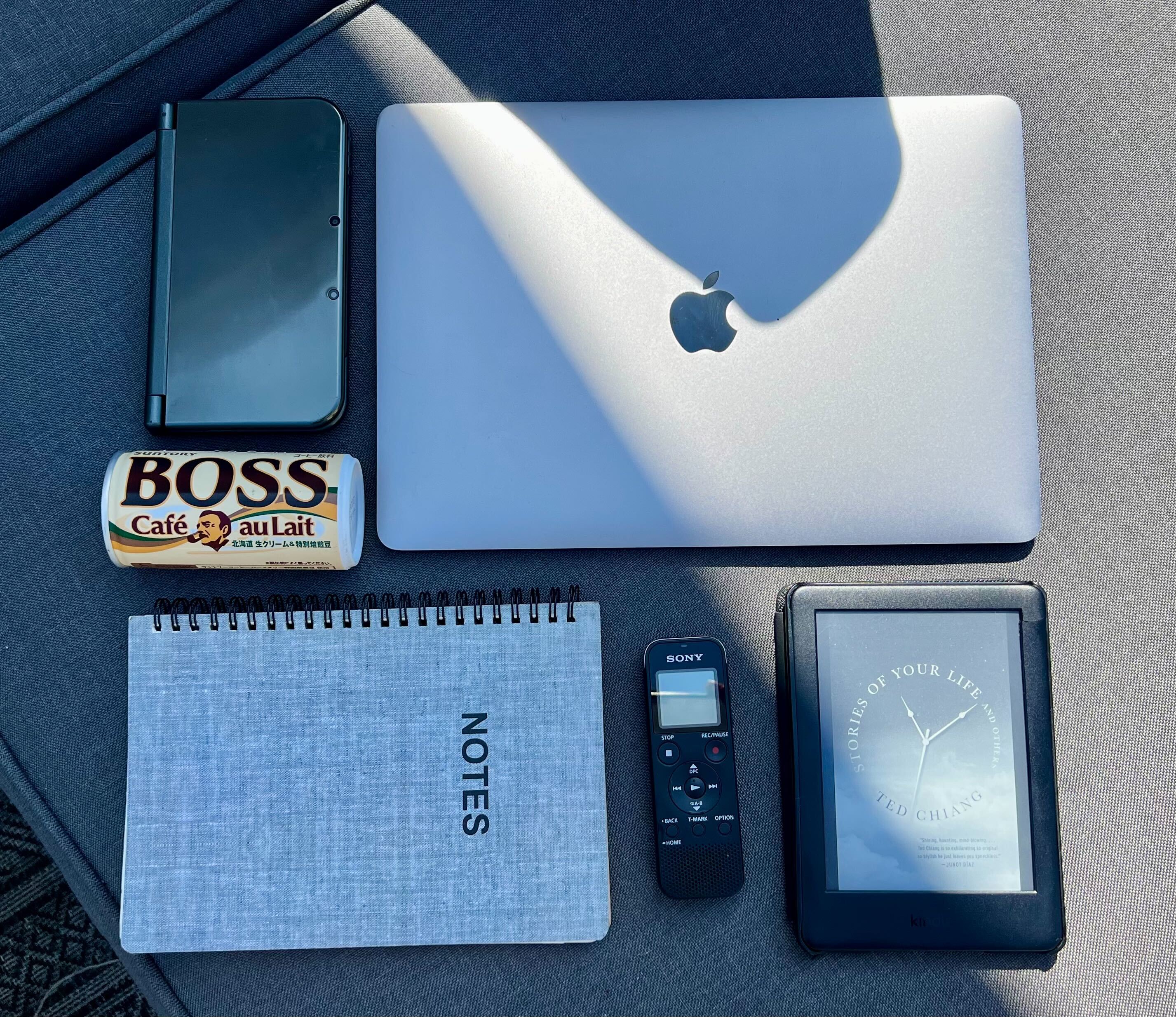 A photo of a laptop, Nintendo 3DS, Kindle, notebook, voice recorder, and can of Boss coffee.