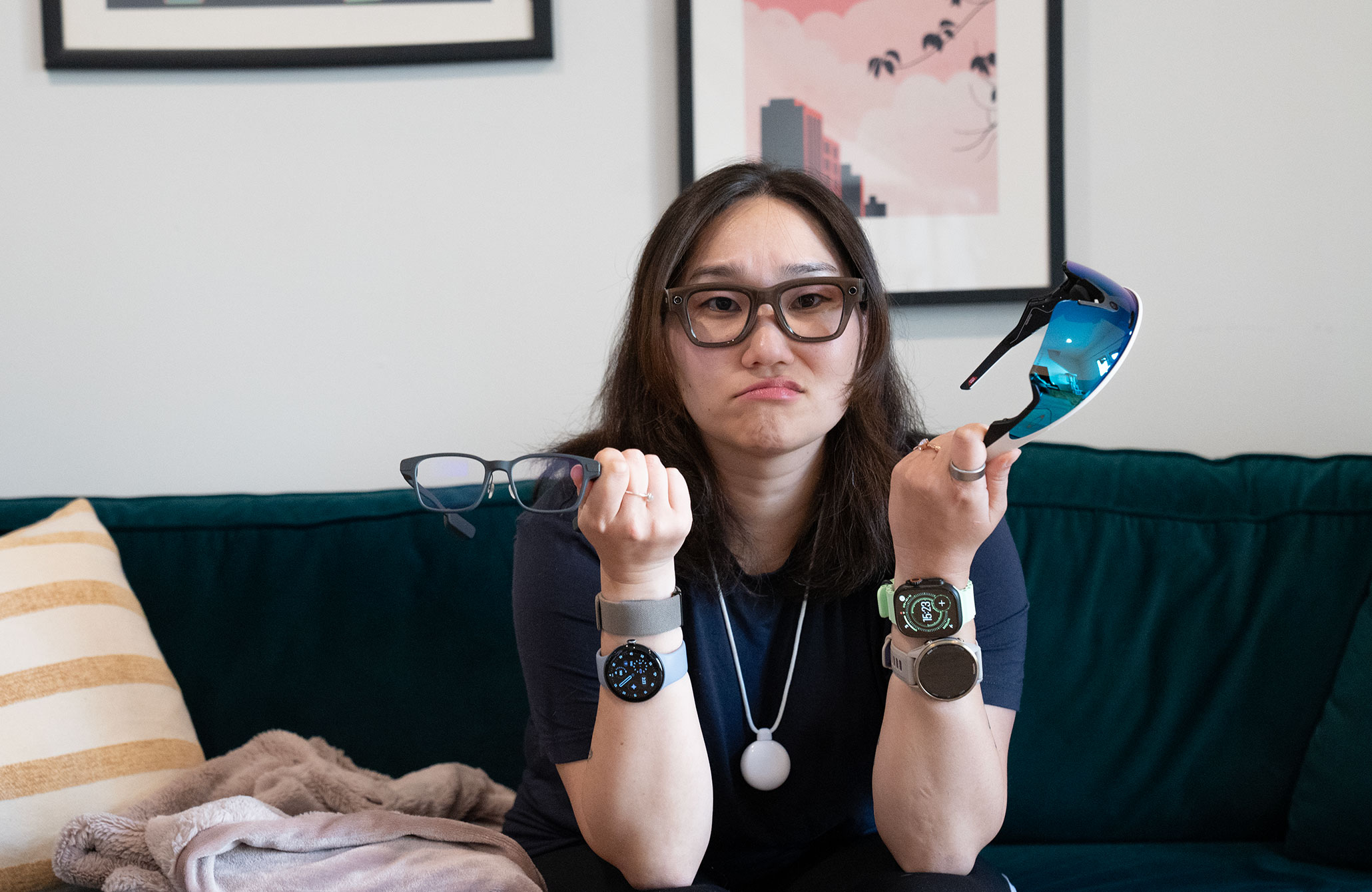 Straight-on view of a disgruntled Senior Reviewer Victoria Song on a green couch wearing a pair of smart glasses, holding two others, and wearing four wrist-based wearables and the Friend AI necklace. On her finger is a smart ring.