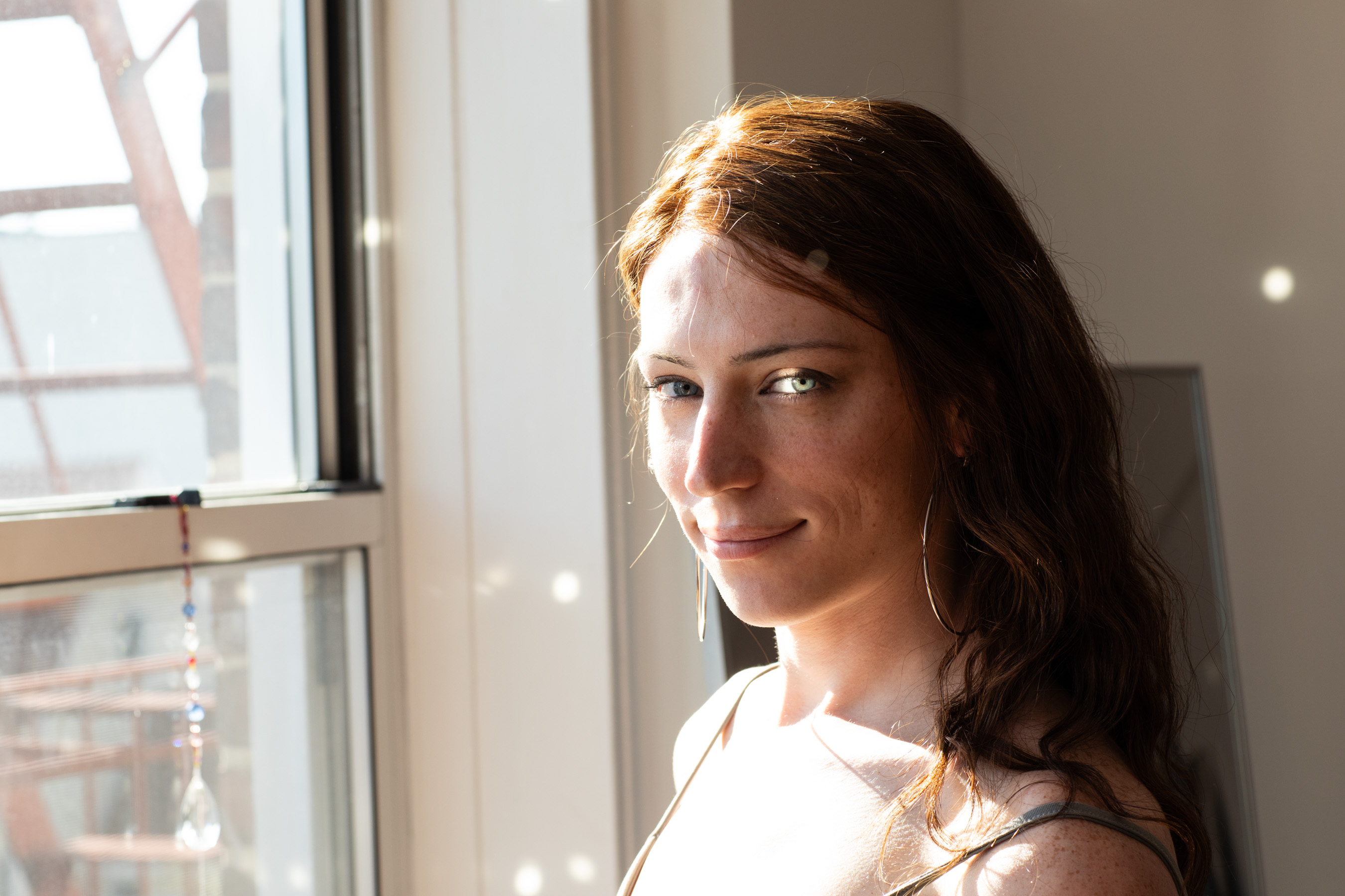 Bree O’Donnell looking at the viewer next to her bedroom window. Sunlight is reflected from a spectrum that bounces little discs of light all over her face and the wall behind her.
