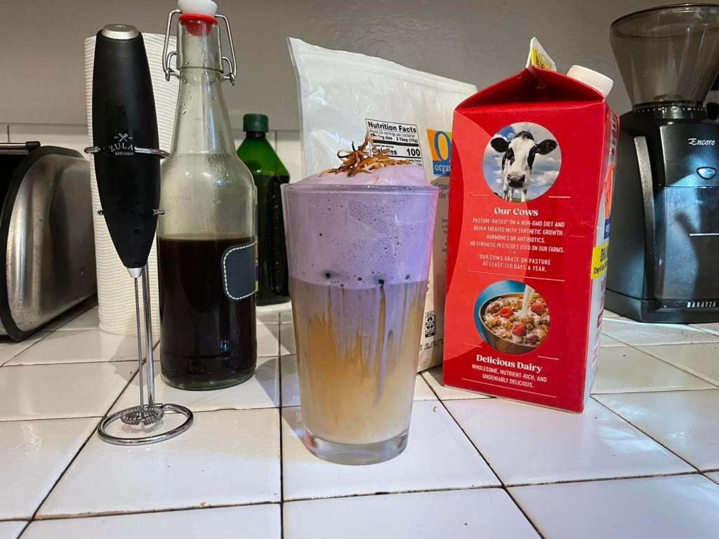 A frothy coffee drink with purple foam and toasted coconut shreds on top, surrounded by the things used to make it: a milk foamer, concentrated iced coffee, coconut shreds, and milk